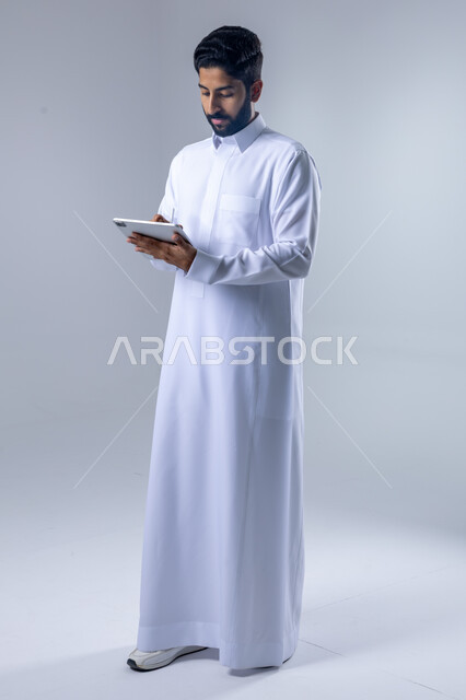 استخدام التقنية الحديثه المتطورة في الحياة العملية عبر الجهاز اللوحي، تعابير وايماءات الاندماج، بورتريه لشاب عربي خليجي سعودي مبتسم يرتدي الثوب التقليدي يقوم بالعمل على جهازالتابلت، صورة الجسم بالطول الكامل، خلفية بيضاء