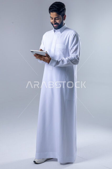Using modern advanced technology in practical life via tablet, expressions and gestures of integration, portrait of a smiling Saudi Arabian Gulf young man wearing traditional dress working on a tablet, full body image, white background