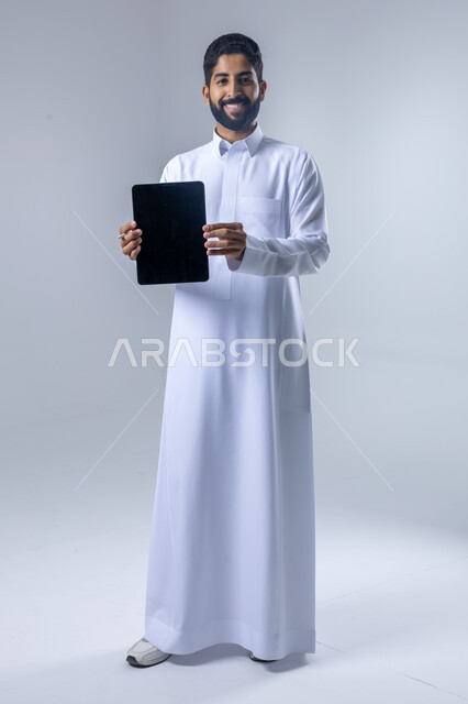 استخدام التقنية الحديثه المتطورة في الحياة العملية عبر الجهاز اللوحي، تعابير وايماءات الاندماج، بورتريه لشاب عربي خليجي سعودي مبتسم يرتدي الثوب التقليدي يعرض شاشة سوداء فارغة على جهازالتابلت، صورة الجسم بالطول الكامل، خلفية بيضاء