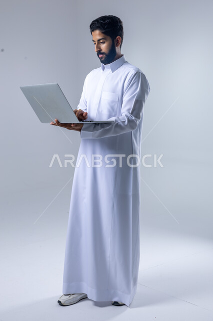 استخدام التقنية الحديثه المتطورة في الحياة العملية، تعابير وايماءات الاندماج والسرور، بورتريه لشاب عربي خليجي سعودي مبتسم يرتدي الثوب التقليدي يقوم بالعمل على الحاسب المحمول، صورة الجسم بالطول الكامل، خلفية بيضاء