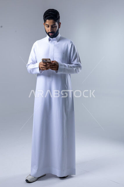  تعابير وايماءات الاندماج والسرور، استخدام التقنية الحديثه المتطورة في الحياة العملية، بورتريه لشاب عربي خليجي سعودي مبتسم يرتدي الثوب التقليدي يقوم بالعمل على الهاتف المحمول، صورة الجسم بالطول الكامل، خلفية بيضاء