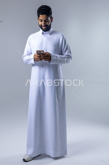  تعابير وايماءات الاندماج والسرور، استخدام التقنية الحديثه المتطورة في الحياة العملية، بورتريه لشاب عربي خليجي سعودي مبتسم يرتدي الثوب التقليدي يقوم بالعمل على الهاتف المحمول، صورة الجسم بالطول الكامل، خلفية بيضاء
