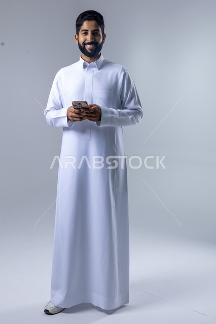  تعابير وايماءات الاندماج والسرور، استخدام التقنية الحديثه المتطورة في الحياة العملية، بورتريه لشاب عربي خليجي سعودي مبتسم يرتدي الثوب التقليدي يقوم بالعمل على الهاتف المحمول، صورة الجسم بالطول الكامل، خلفية بيضاء