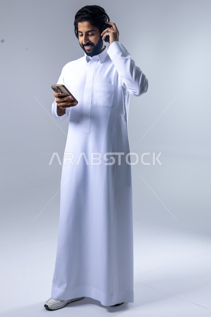 التقنيات والتكنولوجيا الحديثة، بورتريه لشاب عربي سعودي خليجي يرتدي الثوب التقليدي يضع سماعات الهيدفون، التواصل مع الاهل والاقارب عبر الهاتف المحمول، مفهوم الاستماع والاصغاء، الانسجام في سماع الموسيقا والتسجيلات الصوتية، الجسم بالطول الكامل، خلفية بيضاء