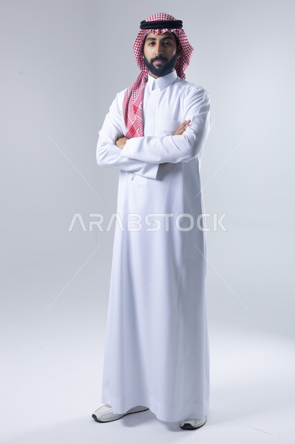 الاعتناء والاهتمام بالمظهر الخارجي، مفهوم الاناقة والرجولة، التقاط صور شخصية للوثائق الرسمية، بورتريه لشاب عربي خليجي سعودي يرتدي الثوب التقليدي والشماغ يقف بأيدي متقاطعة ينظر الى الكاميرا بتعابير السرور والثقة بالنفس، الجسم بالطول الكامل، خلفية بيضاء
