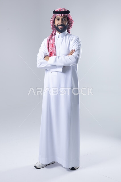 الاعتناء والاهتمام بالمظهر الخارجي، مفهوم الاناقة والرجولة، التقاط صور شخصية للوثائق الرسمية، بورتريه لشاب عربي خليجي سعودي يرتدي الثوب التقليدي والشماغ يقف بأيدي متقاطعة ينظر الى الكاميرا بتعابير السرور والثقة بالنفس، الجسم بالطول الكامل، خلفية بيضاء