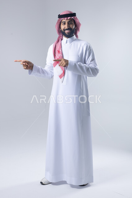 الإشارة إلى شيء ما, الوقوف باستقامة واعتدال, بورتريه لرجل عربي خليجي سعودي يرتدي الثوب التقليدي و الشماغ يشير بيديه لمكان معين, صورة للجسم بالطول الكامل, خلفية بيضاء