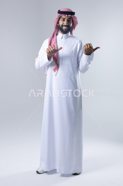 الإشارة إلى شيء ما, الوقوف باستقامة واعتدال, بورتريه لرجل عربي خليجي سعودي يرتدي الثوب التقليدي و الشماغ يشير بيديه لمكان معين, صورة للجسم بالطول الكامل, خلفية بيضاء