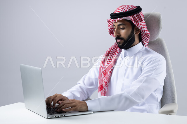The concept of integrating work with technology, completing work tasks via laptop, portrait of a smiling Saudi Arabian Gulf man wearing the traditional thobe and shemagh working on a computer loaded with gestures of pleasure, white background