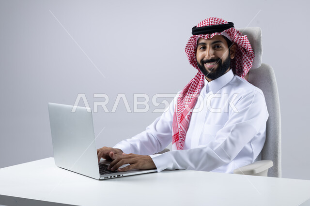 The concept of integrating work with technology, completing work tasks via laptop, portrait of a smiling Saudi Arabian Gulf man wearing the traditional thobe and shemagh working on a computer loaded with gestures of pleasure, white background