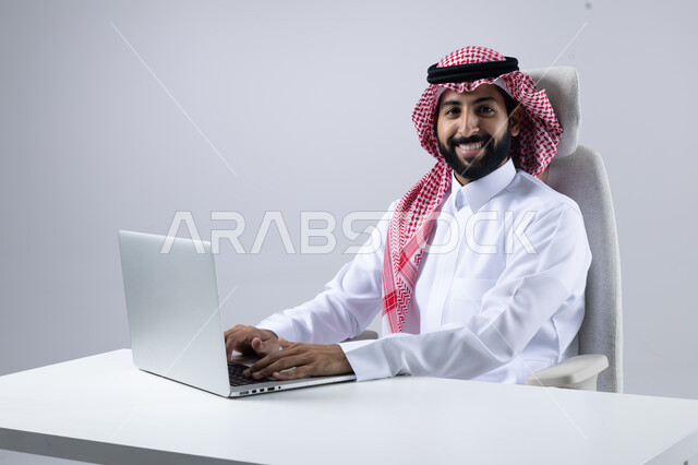 The concept of integrating work with technology, completing work tasks via laptop, portrait of a smiling Saudi Arabian Gulf man wearing the traditional thobe and shemagh working on a computer loaded with gestures of pleasure, white background