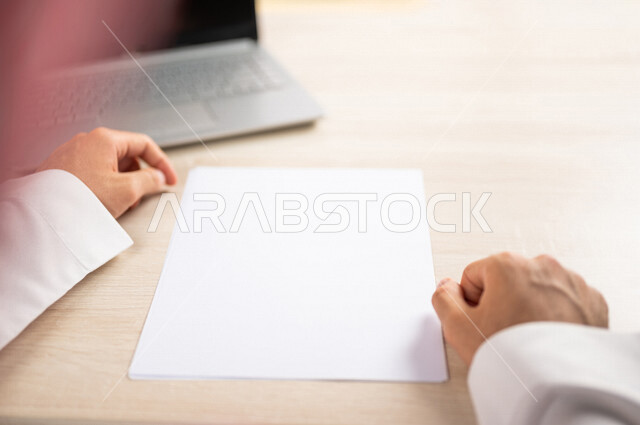 صورة مقربة ليد رجل عربي خليجي سعودي يجلس وأمامه اوراق خاصة بالعمل، أوراق بيضاء فارغة، موظف يجلس أمام الحاسوب المحمول، سكرتير يدير الاعمال الورقية، أعمال مكتبية