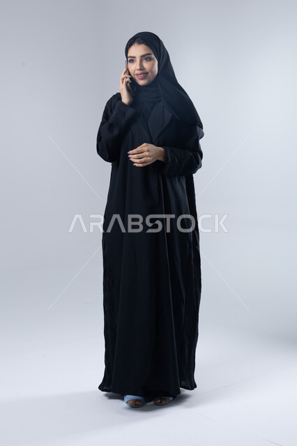 تعابير الاندماج والتركيز بالتحدث عبر الهاتف المحمول، قضاء اوقات ممتعة بتبادل الاخبار والأحاديث، بورتريه لامرأة عربية خليجية سعودية محجبة ترتدي العباءة تقوم بإجرء مكالمة هاتفية، محادثات ودردشات مع الاصدقاء، صورة الجسم بالكامل، خلفية بيضاء