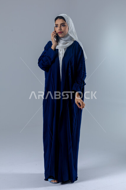 تعابير الاندماج والتركيز بالتحدث عبر الهاتف المحمول، قضاء اوقات ممتعة بتبادل الاخبار والأحاديث، بورتريه لامرأة عربية خليجية سعودية محجبة ترتدي العباءة تقوم بإجرء مكالمة هاتفية، محادثات ودردشات مع الاصدقاء، صورة الجسم بالكامل، خلفية بيضاء