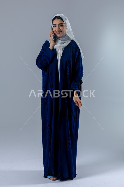 Expressions of immersion and concentration while talking on the mobile phone, having a good time exchanging news and conversations, portrait of a veiled Saudi Arabian Gulf woman wearing an abaya making a phone call, conversations and chats with friends, full body image, white background