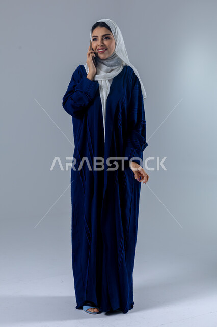 تعابير الاندماج والتركيز بالتحدث عبر الهاتف المحمول، قضاء اوقات ممتعة بتبادل الاخبار والأحاديث، بورتريه لامرأة عربية خليجية سعودية محجبة ترتدي العباءة تقوم بإجرء مكالمة هاتفية، محادثات ودردشات مع الاصدقاء، صورة الجسم بالكامل، خلفية بيضاء