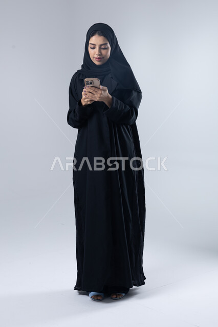  تعابير وايماءات الاندماج والسرور، استخدام التقنية الحديثه المتطورة في الحياة العملية، بورتريه لإمرأة عربية خليجية سعودية ترتدي الحجاب والعباءة تقوم بالعمل على الهاتف المحمول، صورة الجسم بالطول الكامل، خلفية بيضاء 