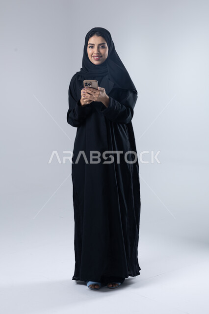  تعابير وايماءات الاندماج والسرور، استخدام التقنية الحديثه المتطورة في الحياة العملية، بورتريه لإمرأة عربية خليجية سعودية ترتدي الحجاب والعباءة تقوم بالعمل على الهاتف المحمول، صورة الجسم بالطول الكامل، خلفية بيضاء 