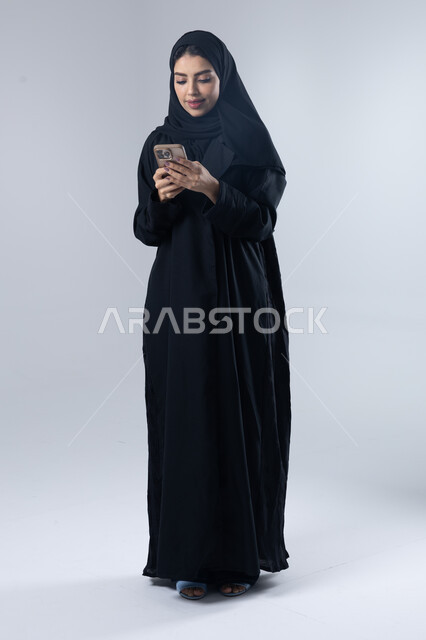  تعابير وايماءات الاندماج والسرور، استخدام التقنية الحديثه المتطورة في الحياة العملية، بورتريه لإمرأة عربية خليجية سعودية ترتدي الحجاب والعباءة تقوم بالعمل على الهاتف المحمول، صورة الجسم بالطول الكامل، خلفية بيضاء 