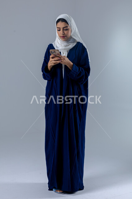  تعابير وايماءات الاندماج والسرور، استخدام التقنية الحديثه المتطورة في الحياة العملية، بورتريه لإمرأة عربية خليجية سعودية ترتدي الحجاب والعباءة تقوم بالعمل على الهاتف المحمول، صورة الجسم بالطول الكامل، خلفية بيضاء 