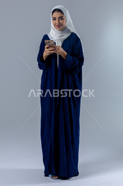  تعابير وايماءات الاندماج والسرور، استخدام التقنية الحديثه المتطورة في الحياة العملية، بورتريه لإمرأة عربية خليجية سعودية ترتدي الحجاب والعباءة تقوم بالعمل على الهاتف المحمول، صورة الجسم بالطول الكامل، خلفية بيضاء 