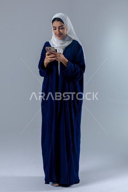  تعابير وايماءات الاندماج والسرور، استخدام التقنية الحديثه المتطورة في الحياة العملية، بورتريه لإمرأة عربية خليجية سعودية ترتدي الحجاب والعباءة تقوم بالعمل على الهاتف المحمول، صورة الجسم بالطول الكامل، خلفية بيضاء 