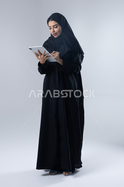  استخدام التقنية الحديثه المتطورة في الحياة العملية عبر الجهاز اللوحي، تعابير وايماءات الاندماج، بورتريه لشابة عربية خليجية سعودية مبتسمة ترتدي الحجاب والعباءة تقوم بالعمل على جهازالتابلت، صورة الجسم بالطول الكامل، خلفية بيضاء 