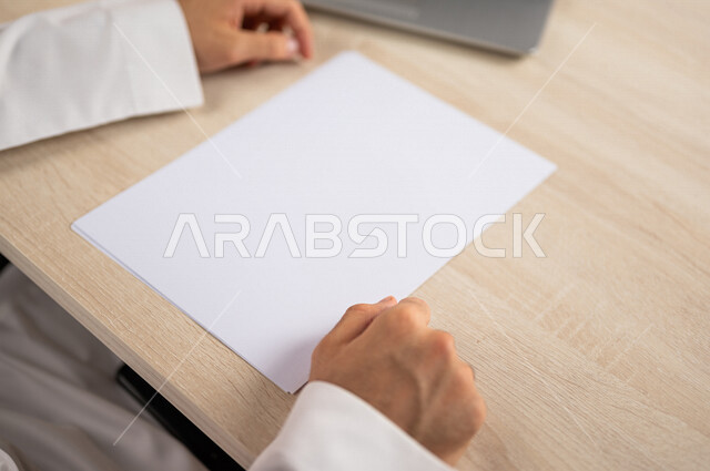 صورة مقربة ليد رجل عربي خليجي سعودي يجلس وأمامه اوراق خاصة بالعمل، أوراق بيضاء فارغة، موظف يجلس أمام الحاسوب المحمول، سكرتير يدير الاعمال الورقية، أعمال مكتبية