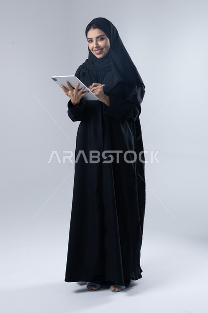  استخدام التقنية الحديثه المتطورة في الحياة العملية عبر الجهاز اللوحي، تعابير وايماءات الاندماج، بورتريه لشابة عربية خليجية سعودية مبتسمة ترتدي الحجاب والعباءة تقوم بالعمل على جهازالتابلت، صورة الجسم بالطول الكامل، خلفية بيضاء 