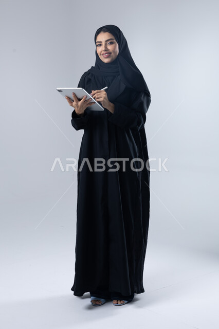 Using modern advanced technology in practical life via tablet, expressions and gestures of integration, portrait of a smiling young Saudi Arabian Gulf woman wearing hijab and abaya working on a tablet, full body image, white background