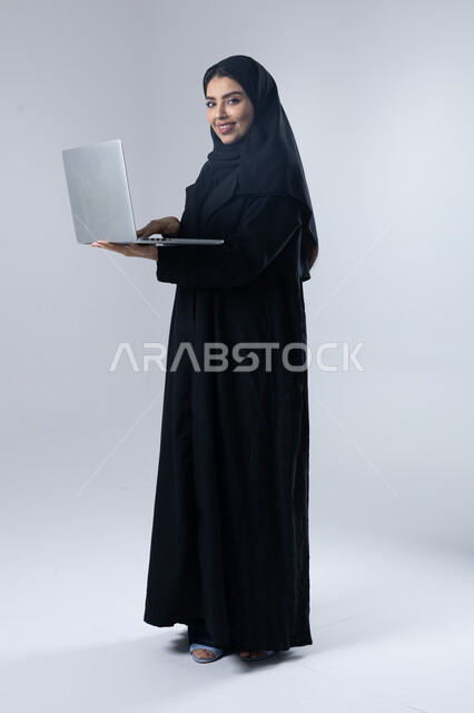  مهن ووظائف نسائية سعودية، ادارة الاعمال عبر التقنية والأجهزة الحديثة، بورتريه لإمرأة عربية خليجية سعودية مبتسمة ترتدي العباءة والحجاب تمسك الحاسب المحمول، التسويق الالكتروني الذكي والآمن، صورة الجسم بالكامل، خلفية بيضاء