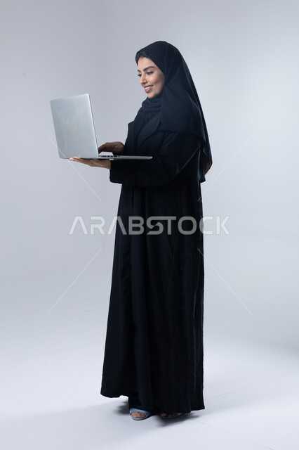  مهن ووظائف نسائية سعودية، ادارة الاعمال عبر التقنية والأجهزة الحديثة، بورتريه لإمرأة عربية خليجية سعودية مبتسمة ترتدي العباءة والحجاب تمسك الحاسب المحمول، التسويق الالكتروني الذكي والآمن، صورة الجسم بالكامل، خلفية بيضاء
