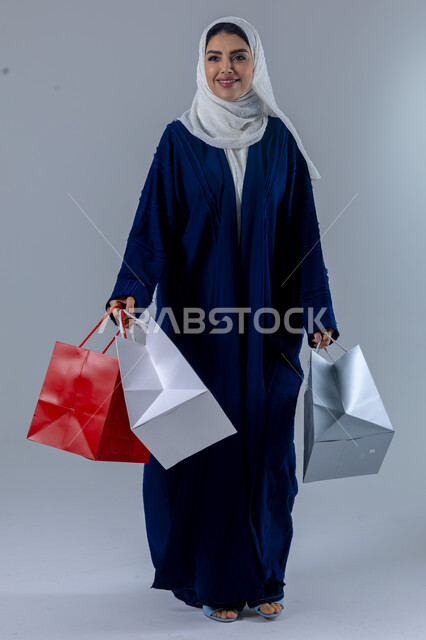 Buying gifts and necessities, taking advantage of seasonal offers and discounts, the positive impact of shopping on women's psychology, portrait of a Saudi Arabian Gulf woman wearing an abaya and hijab, holding colorful bags with gestures of pleasure, full body, white background