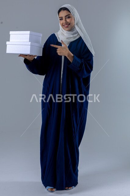 تقديم الهدايا في المناسبات السعيدة, الاحتفال بأعياد الميلاد, التعبير عن المحبة والتقدير, النظر إلى الكاميرا بإيماءات السعادة والسرور, بورتريه لامرأة عربية خليجية سعودية محجبة ترتدي العباءة تمسك بيديها علب هدايا, الجسم بالطول الكامل، خلفية بيضاء