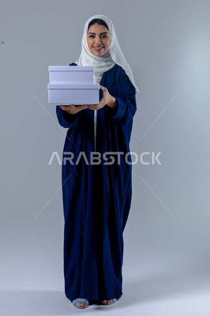 تقديم الهدايا في المناسبات السعيدة, الاحتفال بأعياد الميلاد, التعبير عن المحبة والتقدير, النظر إلى الكاميرا بإيماءات السعادة والسرور, بورتريه لامرأة عربية خليجية سعودية محجبة ترتدي العباءة تمسك بيديها علب هدايا, الجسم بالطول الكامل، خلفية بيضاء