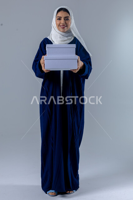 تقديم الهدايا في المناسبات السعيدة, الاحتفال بأعياد الميلاد, التعبير عن المحبة والتقدير, النظر إلى الكاميرا بإيماءات السعادة والسرور, بورتريه لامرأة عربية خليجية سعودية محجبة ترتدي العباءة تمسك بيديها علب هدايا, الجسم بالطول الكامل، خلفية بيضاء