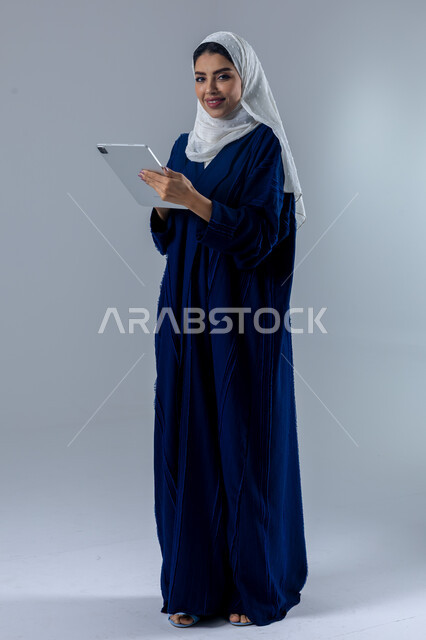 Using modern advanced technology in practical life via tablet, expressions and gestures of integration, portrait of a smiling young Saudi Arabian Gulf woman wearing hijab and abaya working on a tablet, full body image, white background
