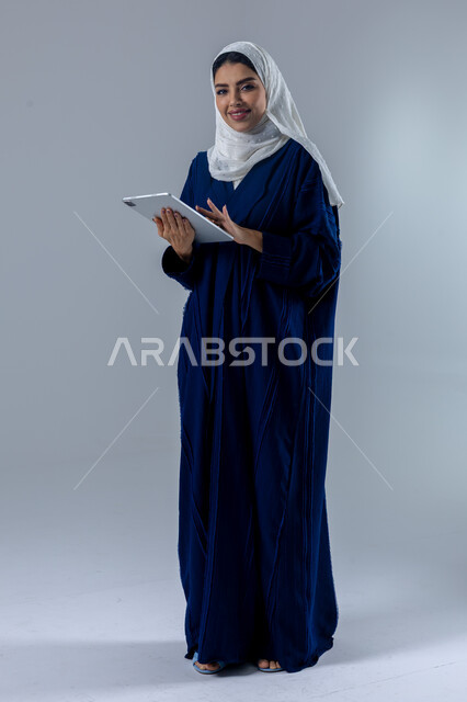  استخدام التقنية الحديثه المتطورة في الحياة العملية عبر الجهاز اللوحي، تعابير وايماءات الاندماج، بورتريه لشابة عربية خليجية سعودية مبتسمة ترتدي الحجاب والعباءة تقوم بالعمل على جهازالتابلت، صورة الجسم بالطول الكامل، خلفية بيضاء 