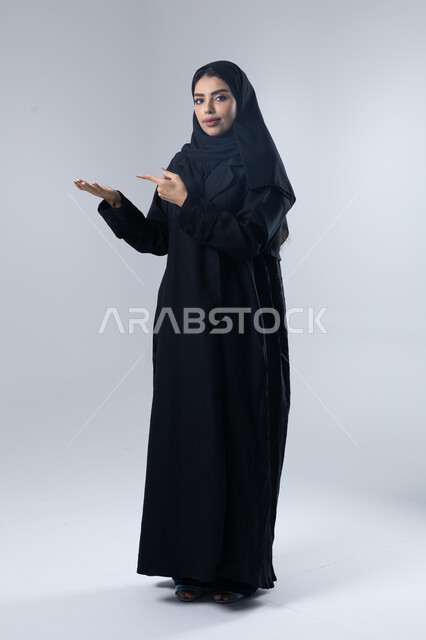 الإشارة إلى عنصر ما, التوضيح والدلالة لمكان محدد, بورتريه لامرأة عربية خليجية سعودية محجبة ترتدي العباءة تشير بيديها على الجانب، تحديد موقع شيء معين, صورة الجسم بالطول الكامل، خلفية بيضاء