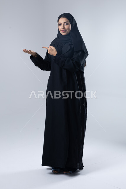 الإشارة إلى عنصر ما, التوضيح والدلالة لمكان محدد, بورتريه لامرأة عربية خليجية سعودية محجبة ترتدي العباءة تشير بيديها على الجانب، تحديد موقع شيء معين, صورة الجسم بالطول الكامل، خلفية بيضاء