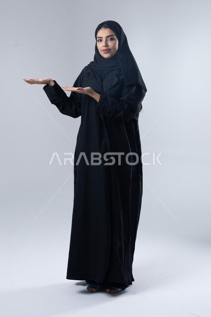 الإشارة إلى عنصر ما, التوضيح والدلالة لمكان محدد, بورتريه لامرأة عربية خليجية سعودية محجبة ترتدي العباءة تشير بيديها على الجانب، تحديد موقع شيء معين, صورة الجسم بالطول الكامل، خلفية بيضاء