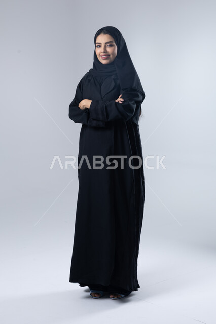 Looking at the camera with gestures of pleasure and self-confidence, the concept of elegance and interest in external appearance, a portrait of a Saudi Arabian Gulf woman wearing the hijab and traditional abaya standing with crossed hands, full-length body image, white background