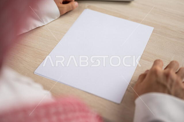 صورة مقربة ليد رجل عربي خليجي سعودي يجلس وأمامه اوراق خاصة بالعمل، أوراق بيضاء فارغة، موظف يجلس أمام الحاسوب المحمول، سكرتير يدير الاعمال الورقية، أعمال مكتبية