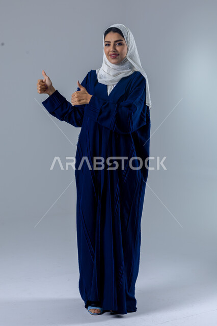 تعابير الموافقة والقبول, النظر الى الكاميرا بإيماءات السرور الرضى والاعجاب، بورتريه لامرأة عربيه خليجية سعودية مبتسمة محجبة ترتدي العباءه تشير باصابع الابهام الى الاعلى بعلامه الامتياز، الطول الكامل للجسم، خلفية بيضاء