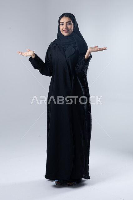 الشعور بالحيرة والجهل بأمر ما، التردد وانعدام المعرفة بخصوص شيء معين، بورتريه لامرأة عربية خليجية سعودية محجبة ترتدي العباءة ترفع يديها بإيماءات رفع عنصر، صورة الجسم بالطول الكامل، خلفية بيضاء