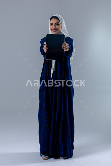 Using modern advanced technology in practical life via tablet, expressions and gestures of integration, portrait of a smiling young Saudi Arabian Gulf woman wearing hijab and abaya displaying a blank black screen on a tablet, full body image, white background