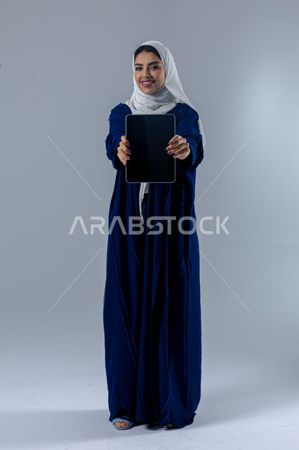  استخدام التقنية الحديثه المتطورة في الحياة العملية عبر الجهاز اللوحي، تعابير وايماءات الاندماج، بورتريه لشابة عربية خليجية سعودية مبتسمة ترتدي الحجاب والعباءة تعرض شاشة سوداء فارغة على جهازالتابلت، صورة الجسم بالطول الكامل، خلفية بيضاء 