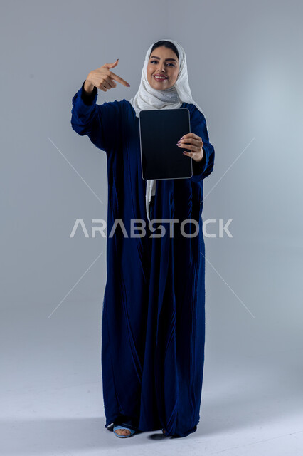 Using modern advanced technology in practical life via tablet, expressions and gestures of integration, portrait of a smiling young Saudi Arabian Gulf woman wearing hijab and abaya displaying a blank black screen on a tablet, full body image, white background