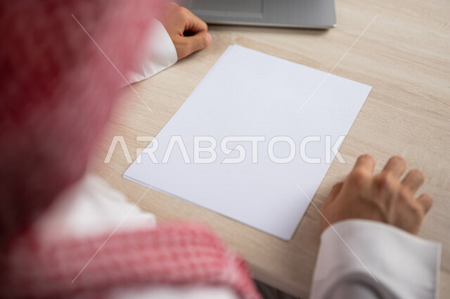 صورة مقربة ليد رجل عربي خليجي سعودي يجلس وأمامه اوراق خاصة بالعمل، أوراق بيضاء فارغة، موظف يجلس أمام الحاسوب المحمول، سكرتير يدير الاعمال الورقية، أعمال مكتبية