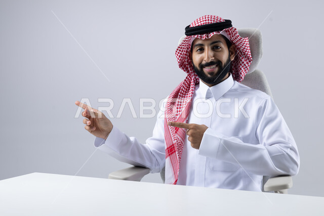 الإشارة إلى شيء ما, الوقوف باستقامة واعتدال, بورتريه مقرب لرجل عربي خليجي سعودي يرتدي الثوب التقليدي و الشماغ يجلس على مكتبه يشير بيديه لمكان معين, خلفية بيضاء